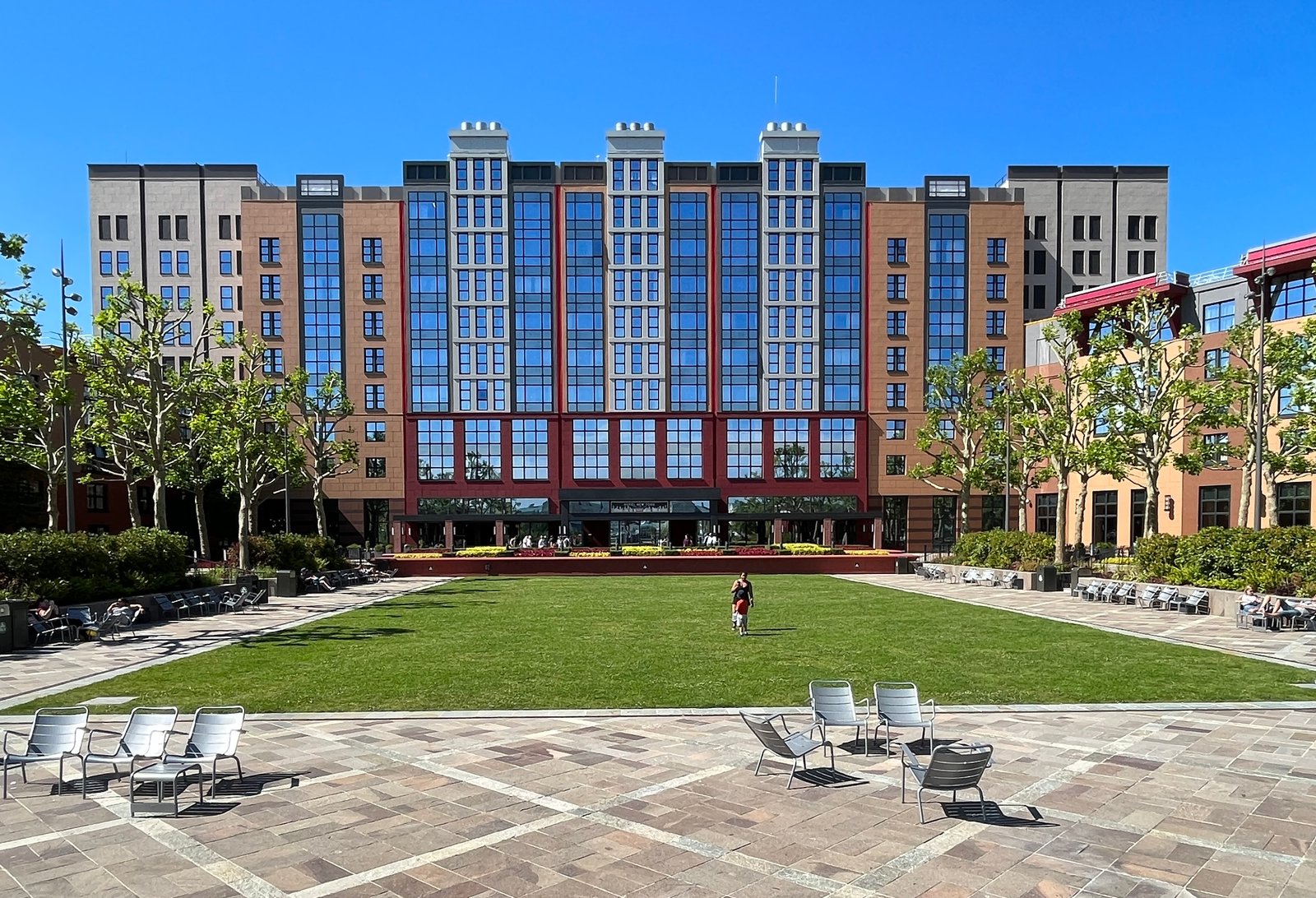 Un grand bâtiment moderne aux hautes baies vitrées, faisant partie d'un Hébergement Disneyland Paris, donne sur une pelouse verte et une cour pavée avec des chaises éparpillées ; une personne seule marche sur l'herbe sous un ciel bleu clair.