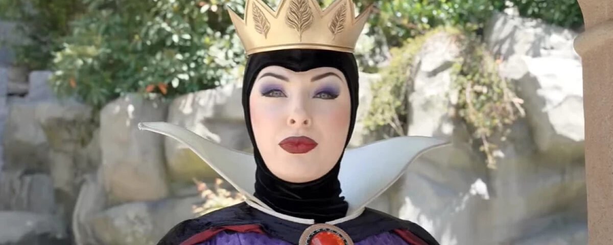 Une personne déguisée en Méchante Reine de Blanche-Neige se tient en plein air à Disneyland, portant une couronne dorée, un maquillage spectaculaire et un costume noir et violet avec un haut col blanc.