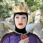 Une personne déguisée en Méchante Reine de Blanche-Neige se tient en plein air à Disneyland, portant une couronne dorée, un maquillage spectaculaire et un costume noir et violet avec un haut col blanc.