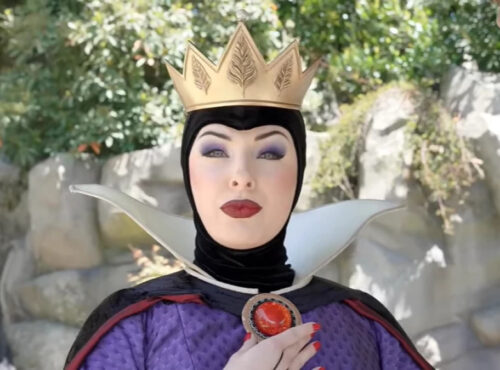 Une personne déguisée en Méchante Reine de Blanche-Neige se tient en plein air à Disneyland, portant une couronne dorée, un maquillage spectaculaire et un costume noir et violet avec un haut col blanc.