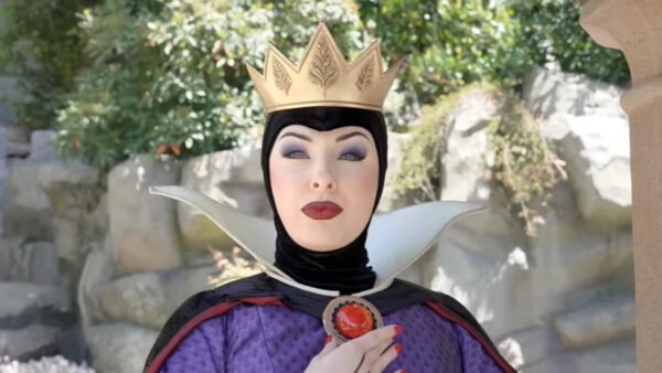 Une personne déguisée en Méchante Reine de Blanche-Neige se tient en plein air à Disneyland, portant une couronne dorée, un maquillage spectaculaire et un costume noir et violet avec un haut col blanc.