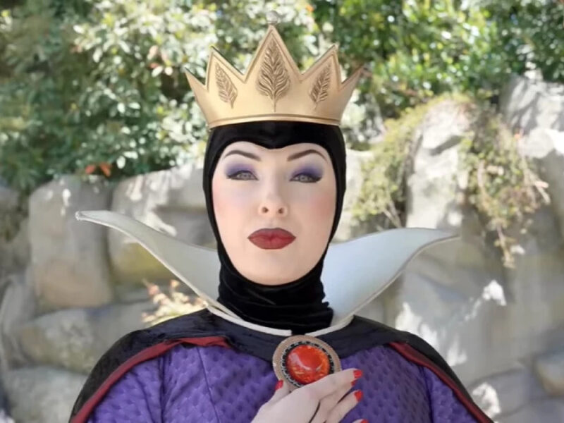 Une personne déguisée en Méchante Reine de Blanche-Neige se tient en plein air à Disneyland, portant une couronne dorée, un maquillage spectaculaire et un costume noir et violet avec un haut col blanc.