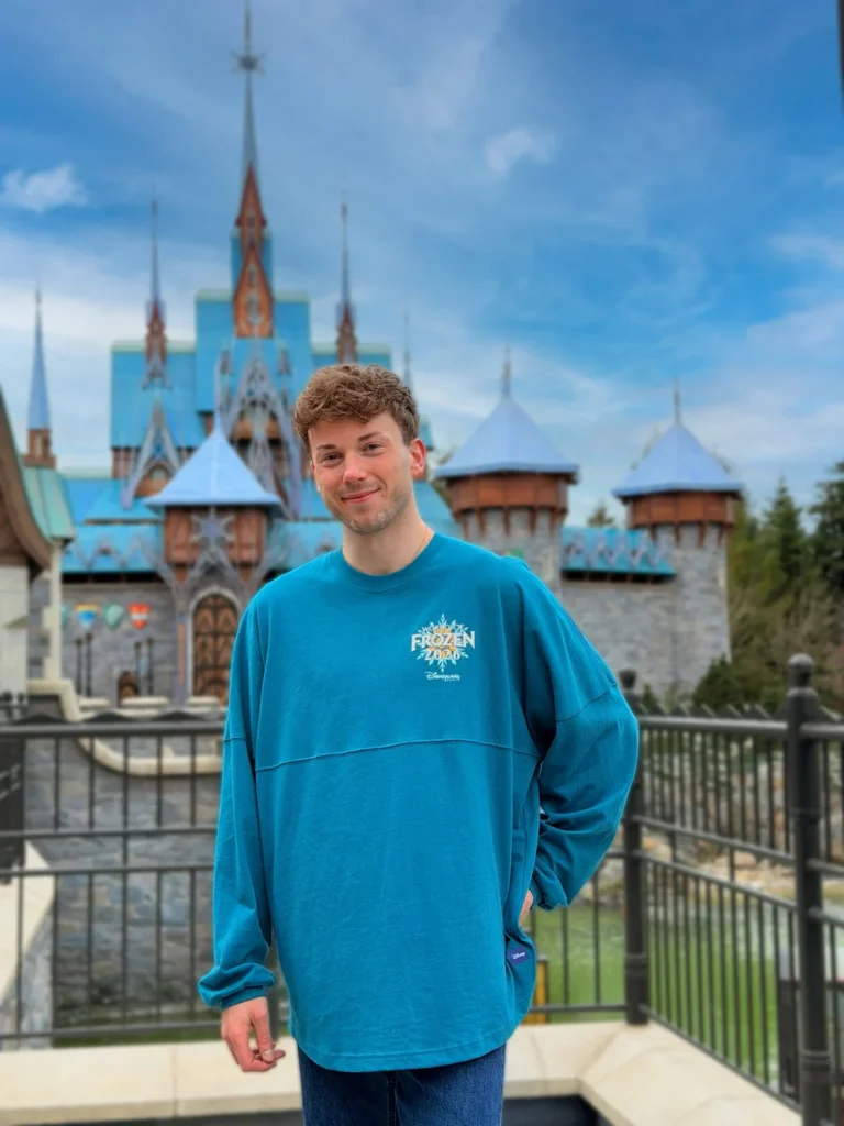 Un jeune homme vêtu d'un sweat-shirt bleu sur le thème de Frozen se tient devant un château aux toits bleus à Disneyland Paris, sous un ciel partiellement nuageux.