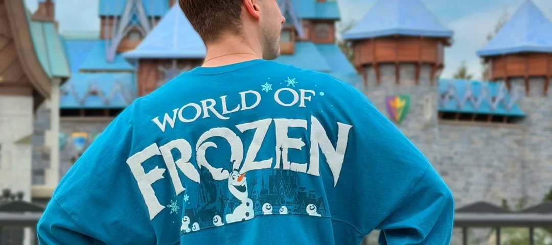 Une personne se tient dos à la caméra, vêtue d'une chemise bleue "World of Frozen", devant un château ressemblant à Arendelle à Disneyland Paris.
