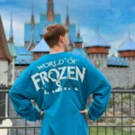 Une personne se tient dos à la caméra, vêtue d'une chemise bleue "World of Frozen", devant un château ressemblant à Arendelle à Disneyland Paris.