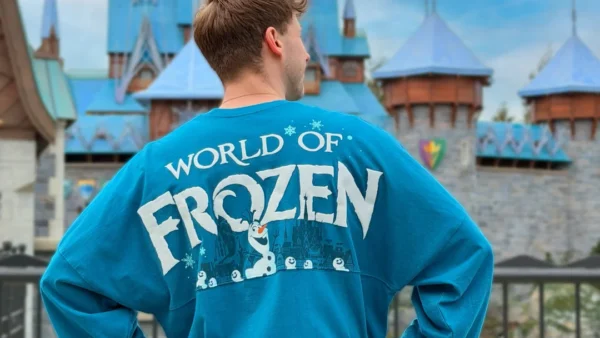 Une personne se tient dos à la caméra, vêtue d'une chemise bleue "World of Frozen", devant un château ressemblant à Arendelle à Disneyland Paris.