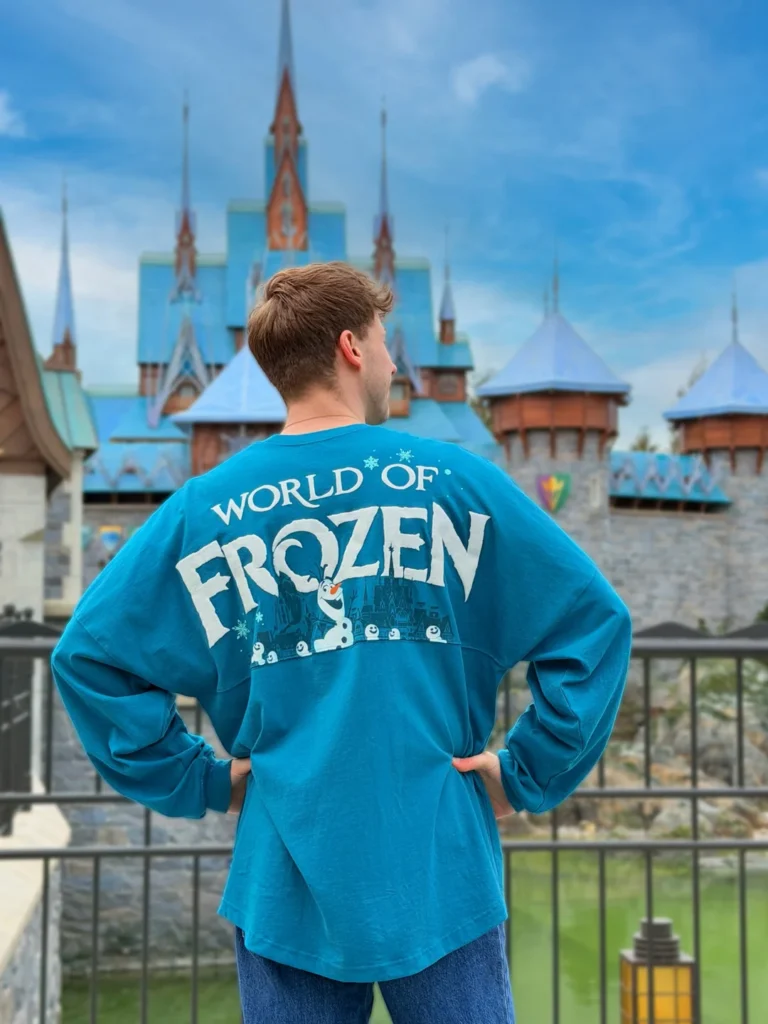 Une personne se tient dos à la caméra, vêtue d'une chemise bleue "World of Frozen", devant un château ressemblant à Arendelle à Disneyland Paris.