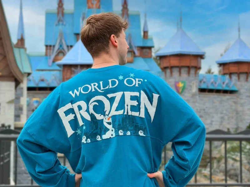 Une personne se tient dos à la caméra, vêtue d'une chemise bleue "World of Frozen", devant un château ressemblant à Arendelle à Disneyland Paris.