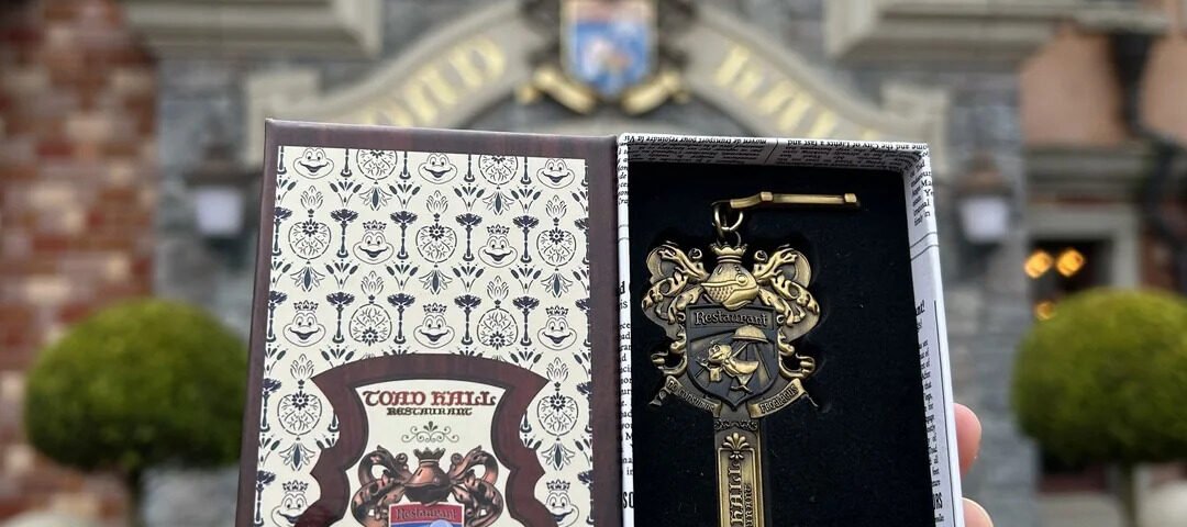 Une main tient une boîte décorative ouverte contenant une clé ornementale dorée, rappelant le manège sauvage de M. Toad à Disneyland, devant un bâtiment en briques dont l'entrée est surmontée d'un écusson.