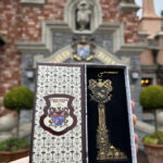 Une main tient une boîte décorative ouverte contenant une clé ornementale dorée, rappelant le manège sauvage de M. Toad à Disneyland, devant un bâtiment en briques dont l'entrée est surmontée d'un écusson.