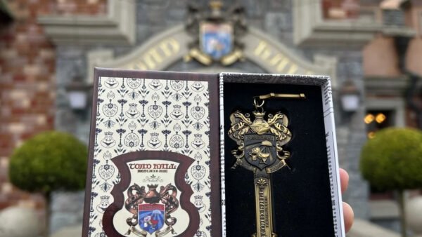 Une main tient une boîte décorative ouverte contenant une clé ornementale dorée, rappelant le manège sauvage de M. Toad à Disneyland, devant un bâtiment en briques dont l'entrée est surmontée d'un écusson.
