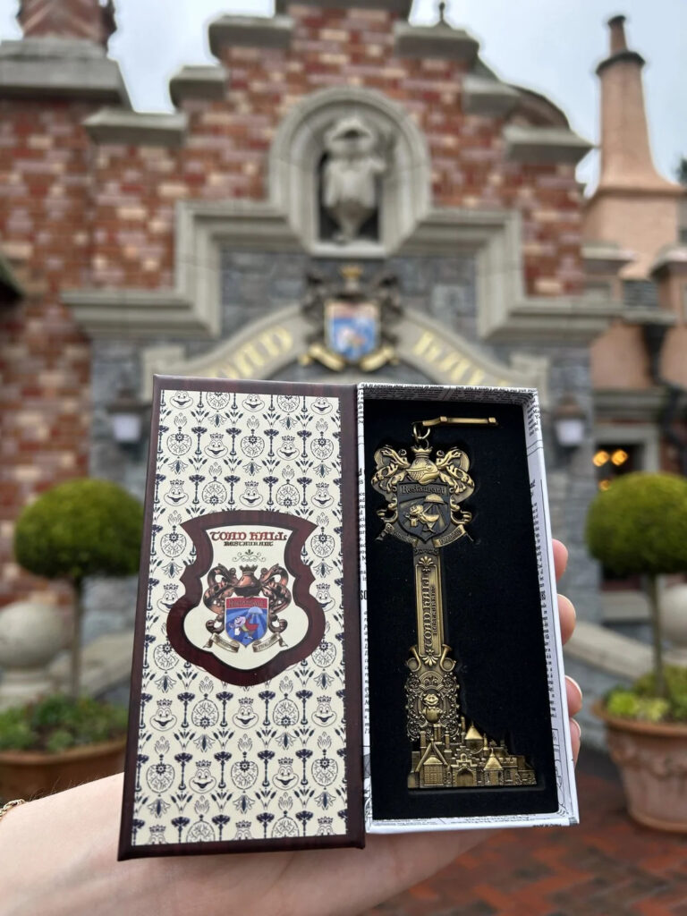 Disneyland. Une main tient une boîte décorative ouverte contenant une clé ornementale dorée, rappelant le manège sauvage de M. Toad à Disneyland, devant un bâtiment en briques dont l'entrée est surmontée d'un écusson.