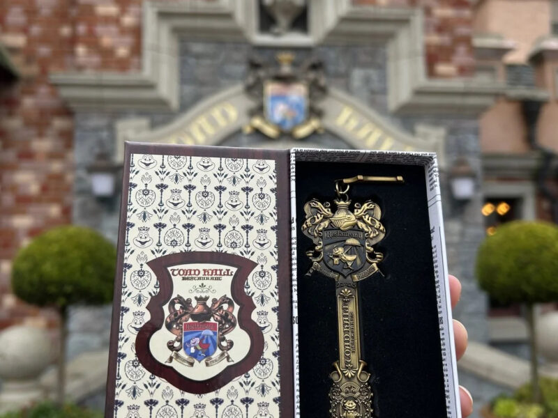 Une main tient une boîte décorative ouverte contenant une clé ornementale dorée, rappelant le manège sauvage de M. Toad à Disneyland, devant un bâtiment en briques dont l'entrée est surmontée d'un écusson.