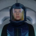 Une personne portant une combinaison de protection bleue et un casque se tient devant une porte circulaire à l'intérieur d'un décor futuriste, rappelant des scènes du film "In The Blink Of An Eye" d'Andrew Stanton, qui sera bientôt disponible sur Disney+.