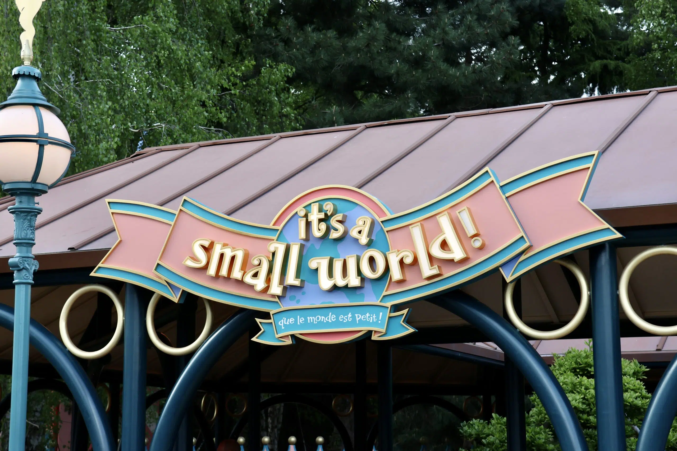 Panneau "it's a small world !" avec traduction en français, monté sur une structure de couleur pastel à Disneyland Paris, entourée d'arbres - l'un des charmants manèges adaptés enfants parfaits pour les familles.