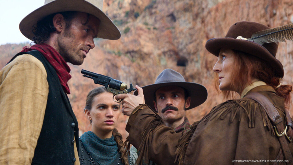 Une femme pointe un revolver sur un homme coiffé d'un chapeau de cow-boy, tandis que deux autres observent la scène. Tous sont vêtus d'habits du vieil Ouest sur fond de rochers, évoquant l'ambiance classique de Lucky Luke que l'on retrouve dans la récente bande-annonce de Disney.