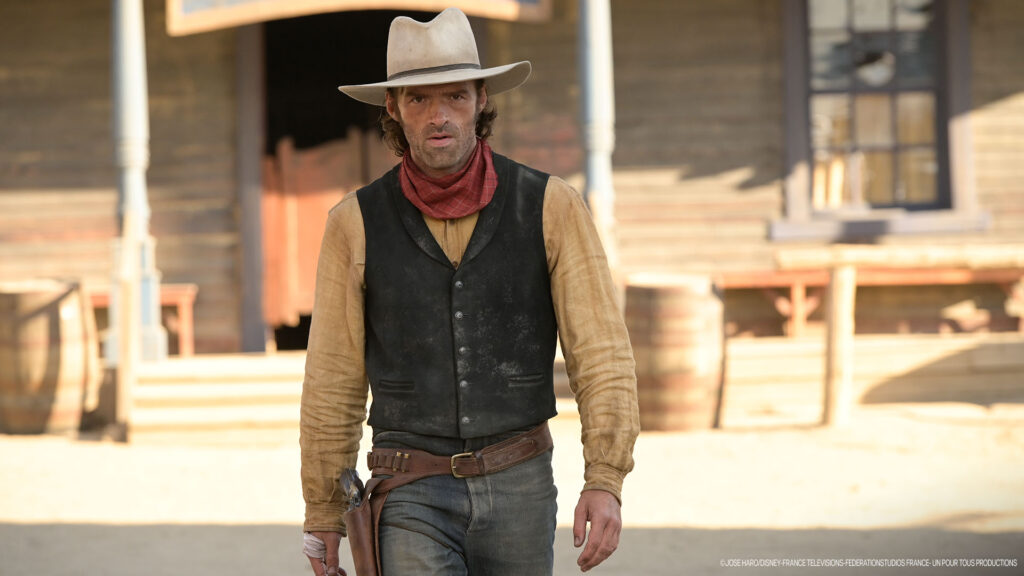 Un homme en tenue de cow-boy, rappelant Lucky Luke, se tient devant un bâtiment en bois dans une ville de style western, avec des barils visibles à l'arrière-plan.