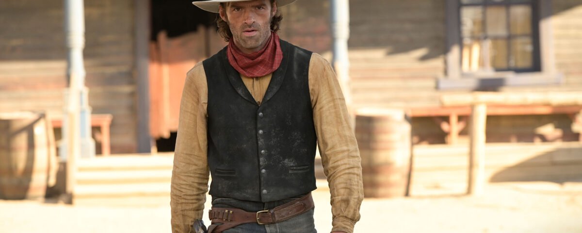 Un homme en tenue de cow-boy, rappelant Lucky Luke, se tient devant un bâtiment en bois dans une ville de style western, avec des barils visibles à l'arrière-plan.