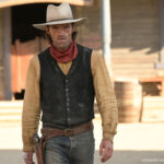 Un homme en tenue de cow-boy, rappelant Lucky Luke, se tient devant un bâtiment en bois dans une ville de style western, avec des barils visibles à l'arrière-plan.