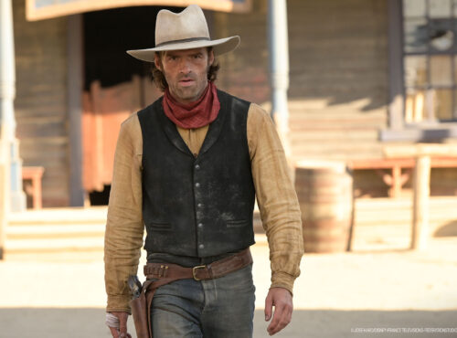 Un homme en tenue de cow-boy, rappelant Lucky Luke, se tient devant un bâtiment en bois dans une ville de style western, avec des barils visibles à l'arrière-plan.