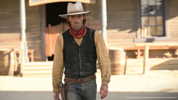 Un homme en tenue de cow-boy, rappelant Lucky Luke, se tient devant un bâtiment en bois dans une ville de style western, avec des barils visibles à l'arrière-plan.