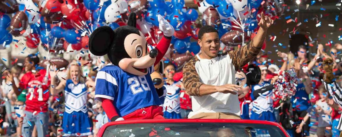 Mickey Mouse en maillot de football et un homme saluent depuis une décapotable rouge lors d'une parade célébrant la tradition de Disneyland avec des pom-pom girls, des ballons et des confettis en arrière-plan.