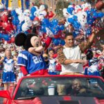 Mickey Mouse en maillot de football et un homme saluent depuis une décapotable rouge lors d'une parade célébrant la tradition de Disneyland avec des pom-pom girls, des ballons et des confettis en arrière-plan.