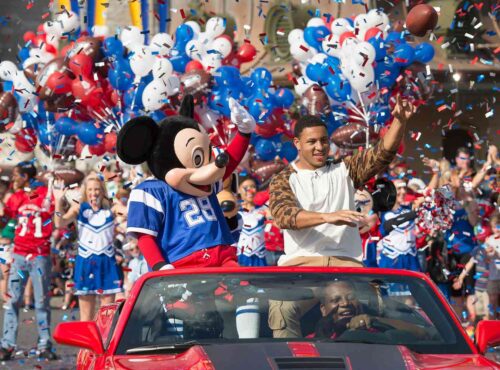 Mickey Mouse en maillot de football et un homme saluent depuis une décapotable rouge lors d'une parade célébrant la tradition de Disneyland avec des pom-pom girls, des ballons et des confettis en arrière-plan.