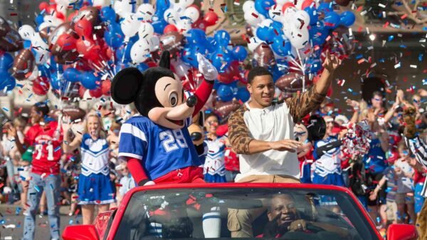 Mickey Mouse en maillot de football et un homme saluent depuis une décapotable rouge lors d'une parade célébrant la tradition de Disneyland avec des pom-pom girls, des ballons et des confettis en arrière-plan.
