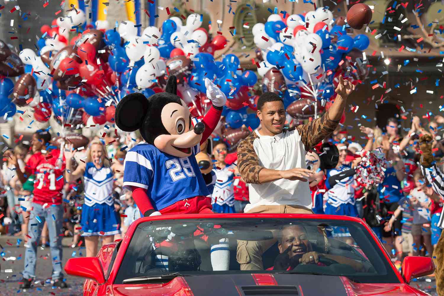 Mickey Mouse en maillot de football et un homme saluent depuis une décapotable rouge lors d'une parade célébrant la tradition de Disneyland avec des pom-pom girls, des ballons et des confettis en arrière-plan.