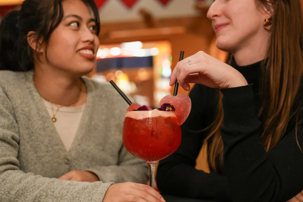 Deux femmes assises à une table de Disneyland Paris se sourient autour d'un grand cocktail rouge agrémenté de fleurs comestibles et d'une rondelle en forme de cœur - un duo de cocktails parfait pour la Saint-Valentin 2026.