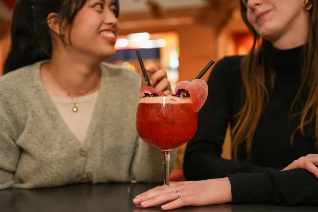 Deux femmes sont assises à une table de Disneyland Paris, souriantes et discutant, avec un grand cocktail rouge de la carte Cocktails Duo Exclusifs, agrémenté de fleurs et de décorations en forme de cœur, parfait pour fêter la Saint-Valentin 2026.