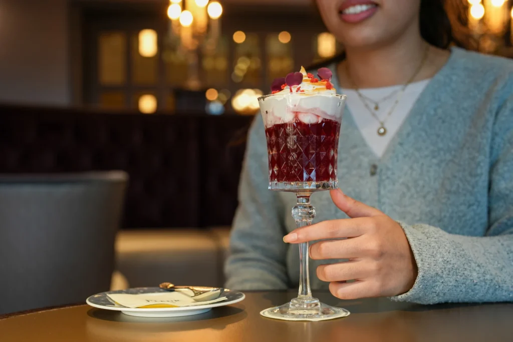 Une personne tient un verre décoratif rempli d'un dessert en couches garni de crème fouettée et de pétales de fleurs, assise à une table avec une assiette, une fourchette, et dégustant des cocktails duo exclusifs inspirés de Disneyland Paris.