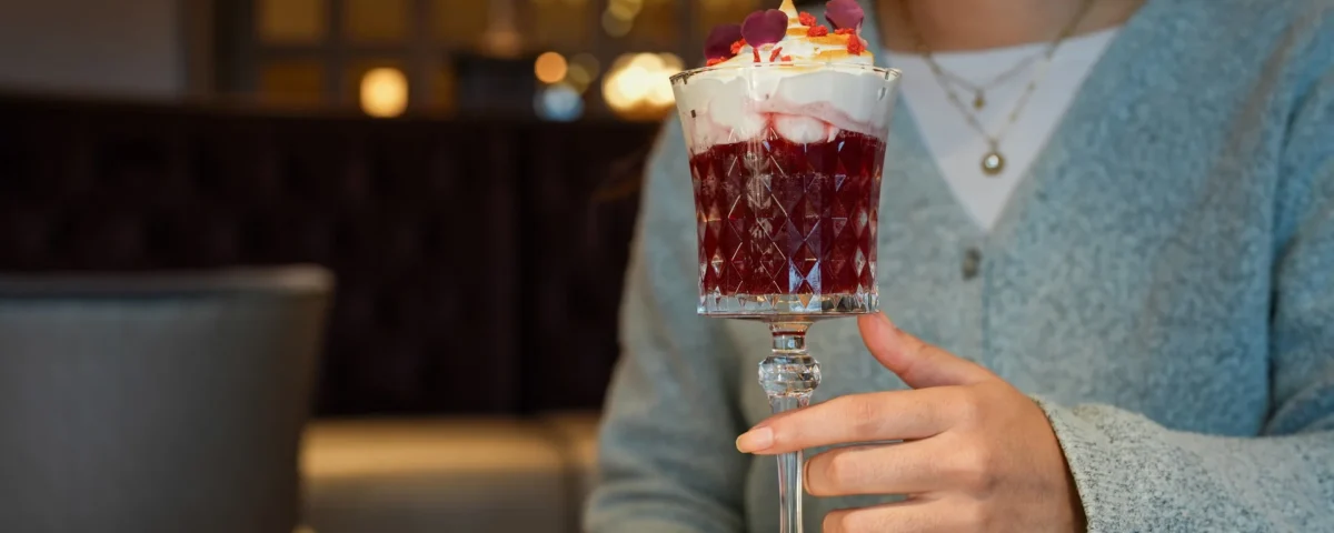 Une personne tient un verre décoratif rempli d'un dessert en couches garni de crème fouettée et de pétales de fleurs, assise à une table avec une assiette, une fourchette, et dégustant des cocktails duo exclusifs inspirés de Disneyland Paris.