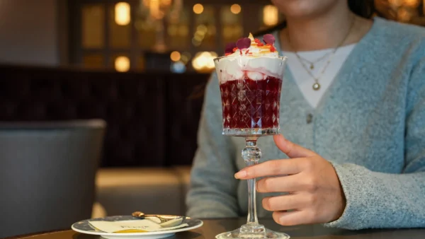 Une personne tient un verre décoratif rempli d'un dessert en couches garni de crème fouettée et de pétales de fleurs, assise à une table avec une assiette, une fourchette, et dégustant des cocktails duo exclusifs inspirés de Disneyland Paris.