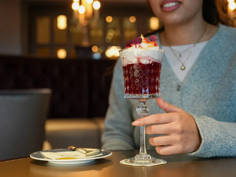 Une personne tient un verre décoratif rempli d'un dessert en couches garni de crème fouettée et de pétales de fleurs, assise à une table avec une assiette, une fourchette, et dégustant des cocktails duo exclusifs inspirés de Disneyland Paris.
