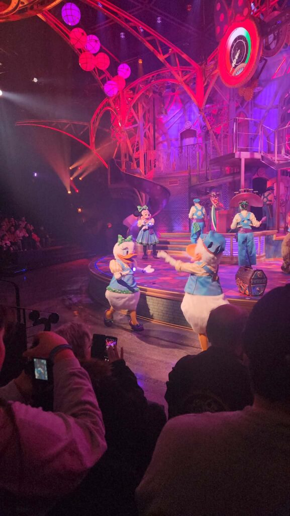 Les personnages Disney de la Fabrique Des Rêves De Minnie se produisent sur une scène colorée et illuminée au Studio D de Disneyland Paris, tandis que le public observe et prend des photos au premier plan.