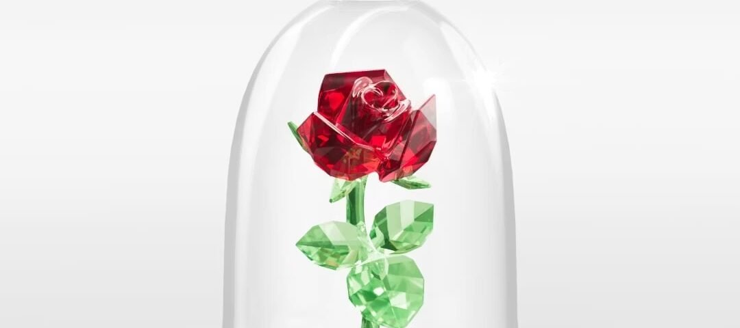 Une rose en cristal rouge et vert de Swarovski est présentée sous un dôme en verre transparent, rappelant La Belle et la Bête, sur un fond blanc uni.