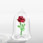 Une rose en cristal rouge et vert de Swarovski est présentée sous un dôme en verre transparent, rappelant La Belle et la Bête, sur un fond blanc uni.