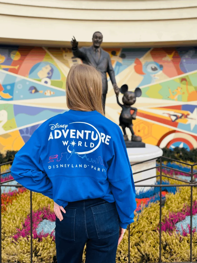 Une personne se tient debout, dos à la caméra, vêtue d'un sweat-shirt bleu de Disney Adventure World, devant une statue de Walt Disney et de Mickey Mouse et une fresque murale colorée à Disneyland Paris.