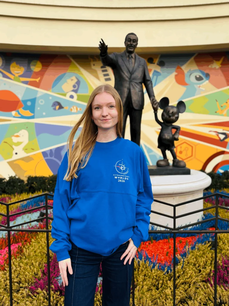 Une femme vêtue d'un sweat-shirt bleu se tient devant la statue des partenaires de Walt Disney et de Mickey Mouse à Disneyland Paris, avec une peinture murale colorée et des fleurs en arrière-plan.