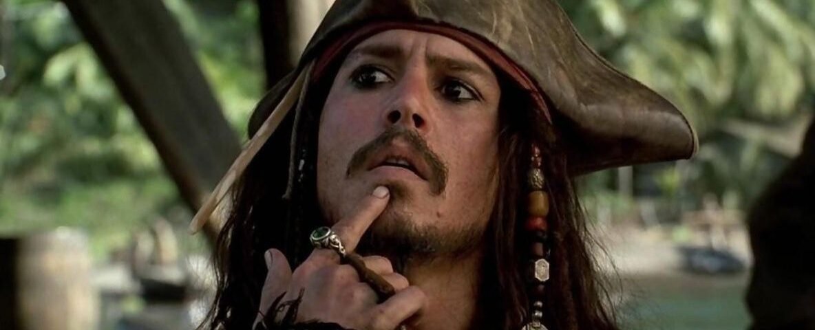 Un homme habillé en pirate avec un chapeau tricorne et des perles dans les cheveux se touche le menton, l'air pensif ; serait-il en train de réfléchir au reboot de "Pirates des Caraïbes 6" par le scénariste de 1917 ? Des palmiers se balancent en arrière-plan.