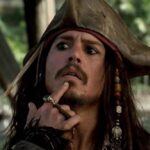 Un homme habillé en pirate avec un chapeau tricorne et des perles dans les cheveux se touche le menton, l'air pensif ; serait-il en train de réfléchir au reboot de "Pirates des Caraïbes 6" par le scénariste de 1917 ? Des palmiers se balancent en arrière-plan.