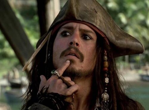 Un homme habillé en pirate avec un chapeau tricorne et des perles dans les cheveux se touche le menton, l'air pensif ; serait-il en train de réfléchir au reboot de "Pirates des Caraïbes 6" par le scénariste de 1917 ? Des palmiers se balancent en arrière-plan.