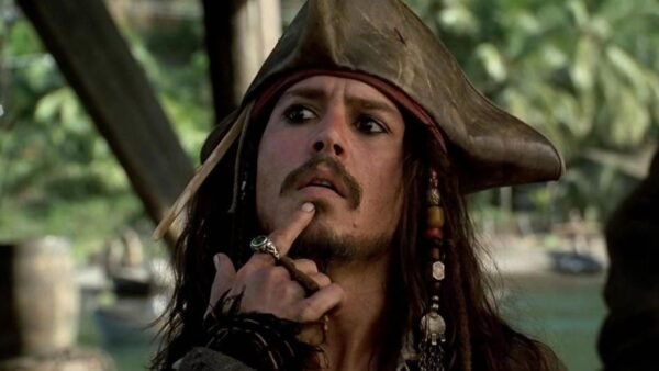 Un homme habillé en pirate avec un chapeau tricorne et des perles dans les cheveux se touche le menton, l'air pensif ; serait-il en train de réfléchir au reboot de "Pirates des Caraïbes 6" par le scénariste de 1917 ? Des palmiers se balancent en arrière-plan.