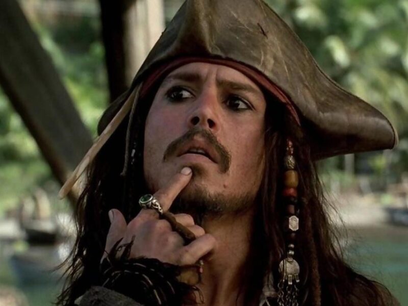 Un homme habillé en pirate avec un chapeau tricorne et des perles dans les cheveux se touche le menton, l'air pensif ; serait-il en train de réfléchir au reboot de "Pirates des Caraïbes 6" par le scénariste de 1917 ? Des palmiers se balancent en arrière-plan.