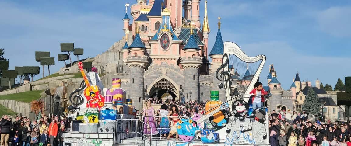 Un char de parade avec des personnages de Disney Pixar passe devant le château de la Belle au bois dormant à Disneyland Paris, sous le regard de la foule dans un ciel bleu clair.