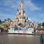 Un char de parade avec des personnages de Disney Pixar passe devant le château de la Belle au bois dormant à Disneyland Paris, sous le regard de la foule dans un ciel bleu clair.