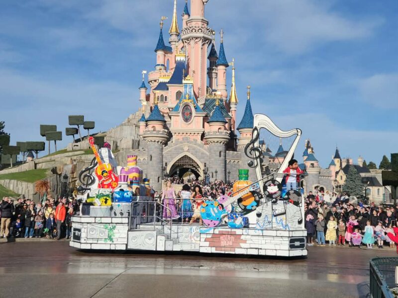 Un char de parade avec des personnages de Disney Pixar passe devant le château de la Belle au bois dormant à Disneyland Paris, sous le regard de la foule dans un ciel bleu clair.
