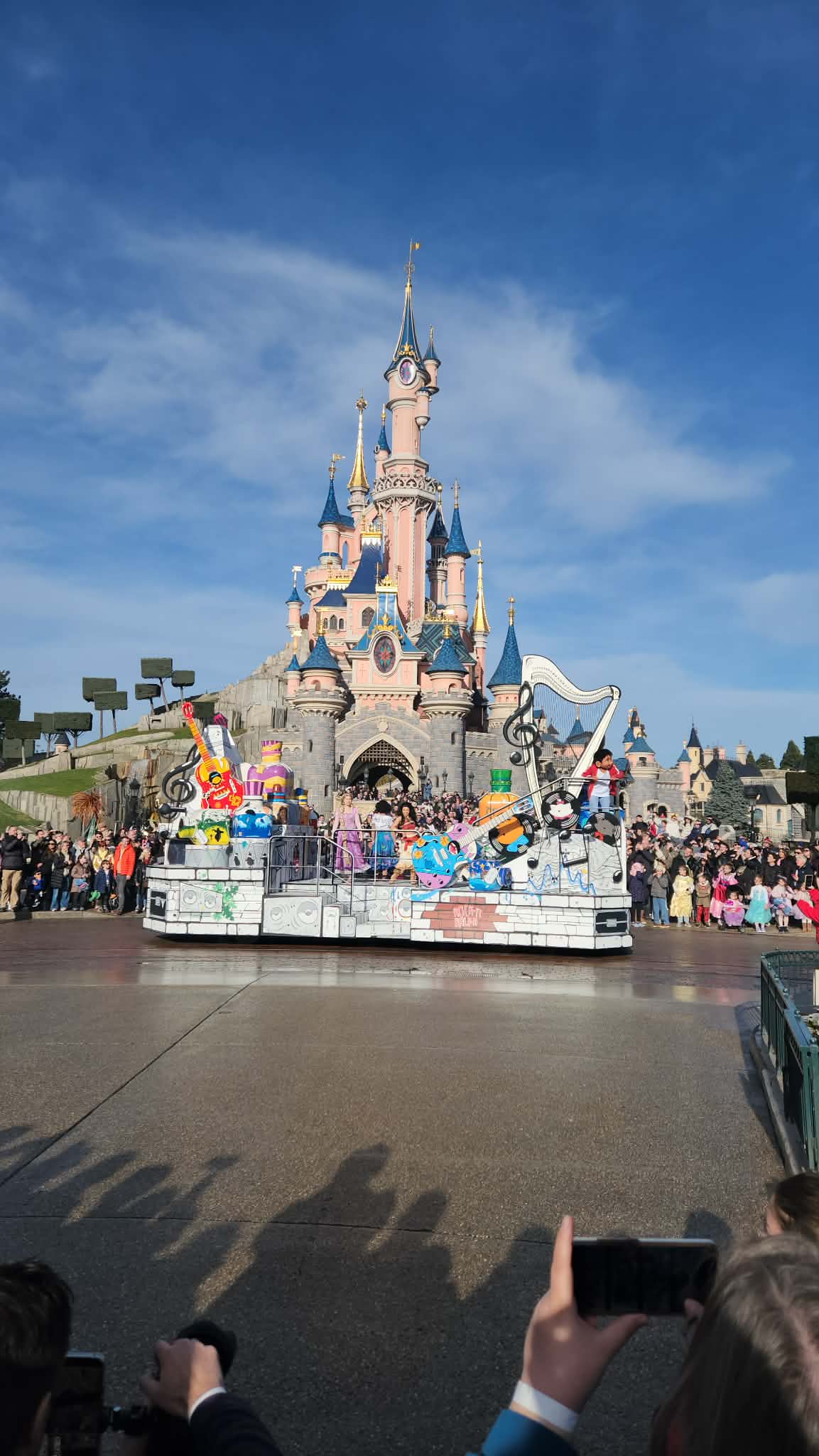 Un char de parade avec des personnages de Disney Pixar passe devant le château de la Belle au bois dormant à Disneyland Paris, sous le regard de la foule dans un ciel bleu clair.